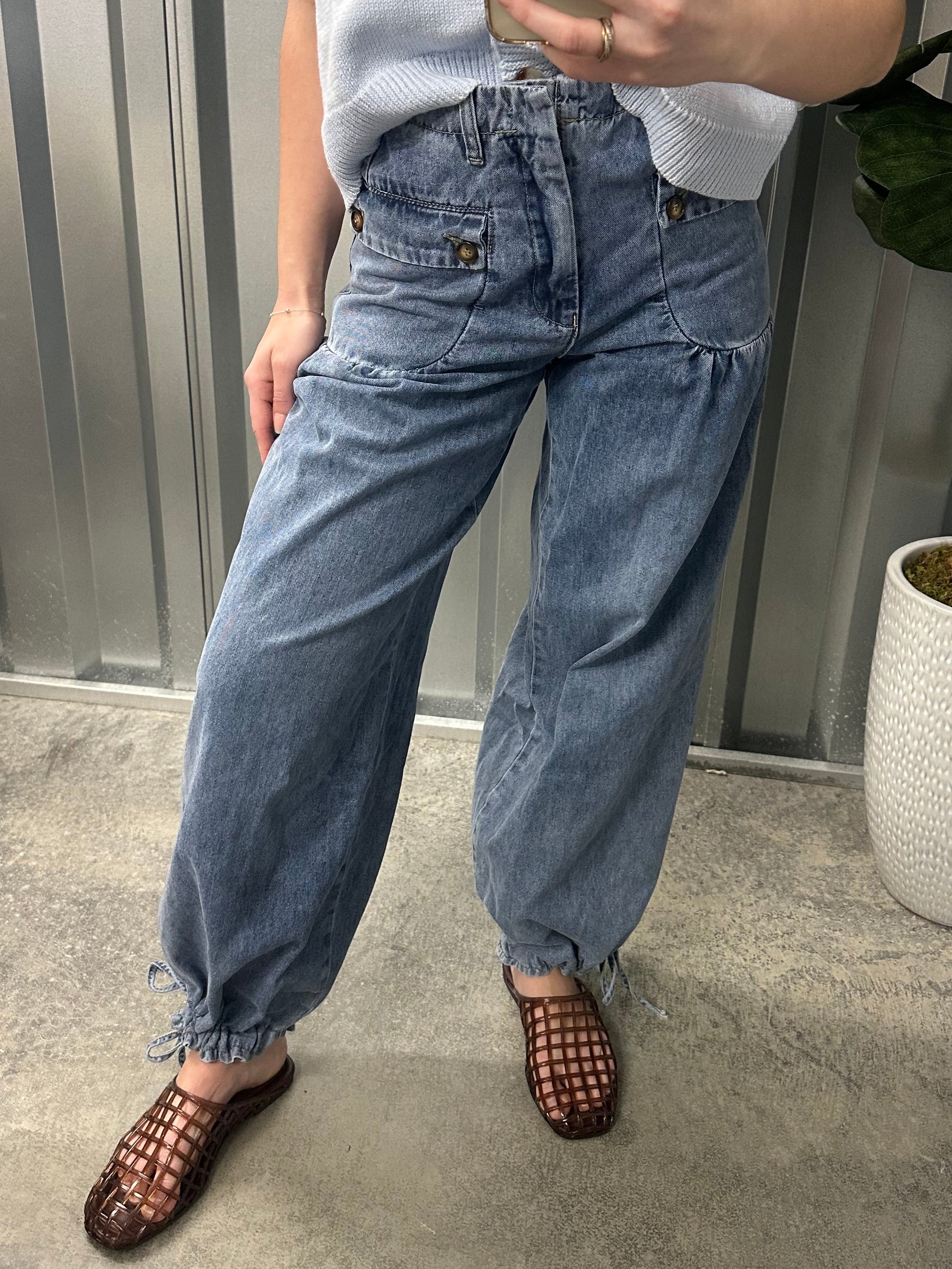 Person taking a mirror selfie wearing a white shirt, blue jeans, and sandals on a concrete floor.