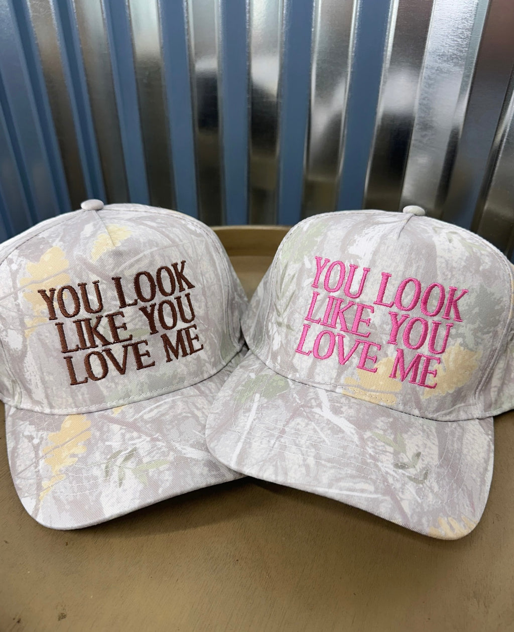 Two baseball caps with text 'YOU LOOK LIKE YOU LOVE ME' in different colors on a metallic background.