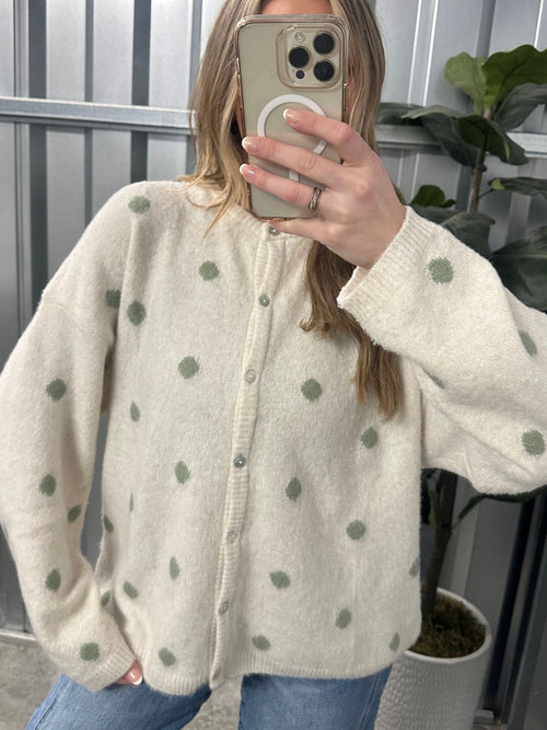 Person wearing a cream-colored cardigan with green polka dots, taking a mirror selfie.
