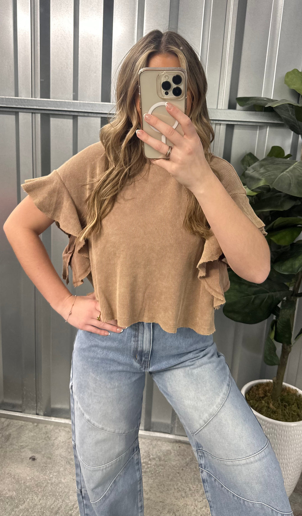 Person wearing a beige top and blue jeans standing outdoors with a plant in the background
