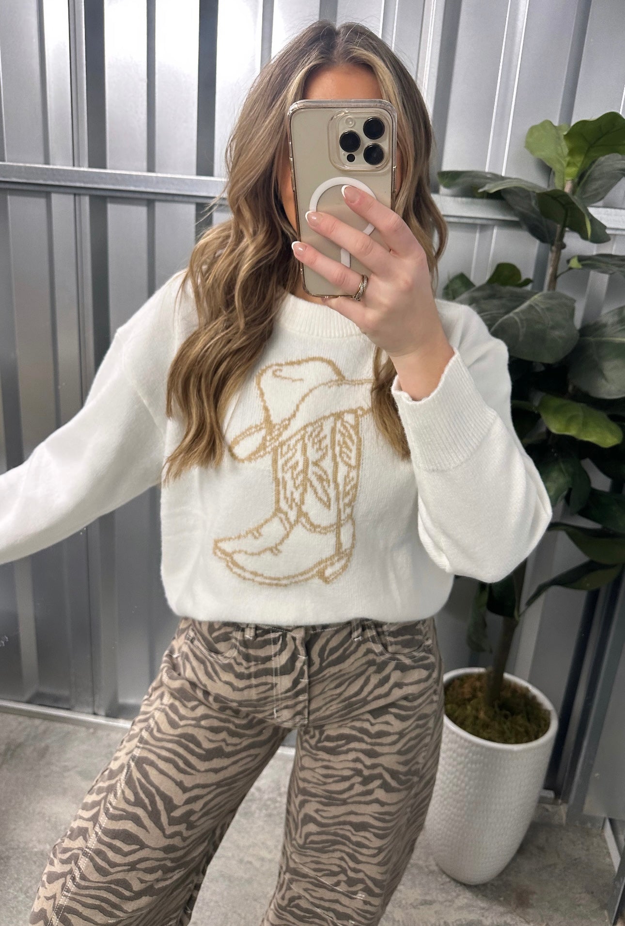 Person wearing a white sweater with a gold boot design and tiger print pants, taking a mirror selfie.